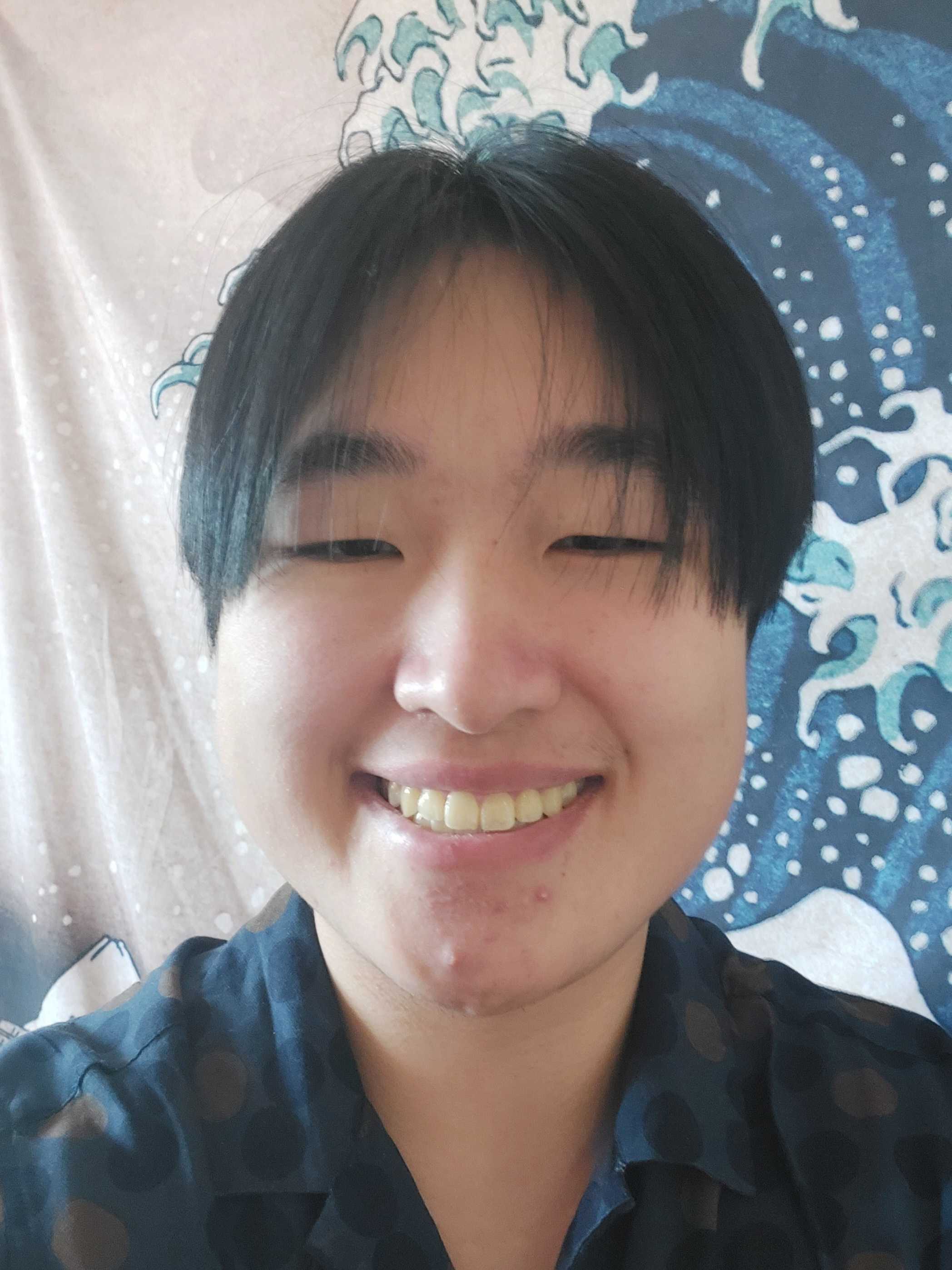 An image of Kelly Wang, who has short, straight black hair down to their cheekbones and is wearing a dark blue collared shirt. He is smiling in front of a tapestry of the Great Wave Off Kanagawa.